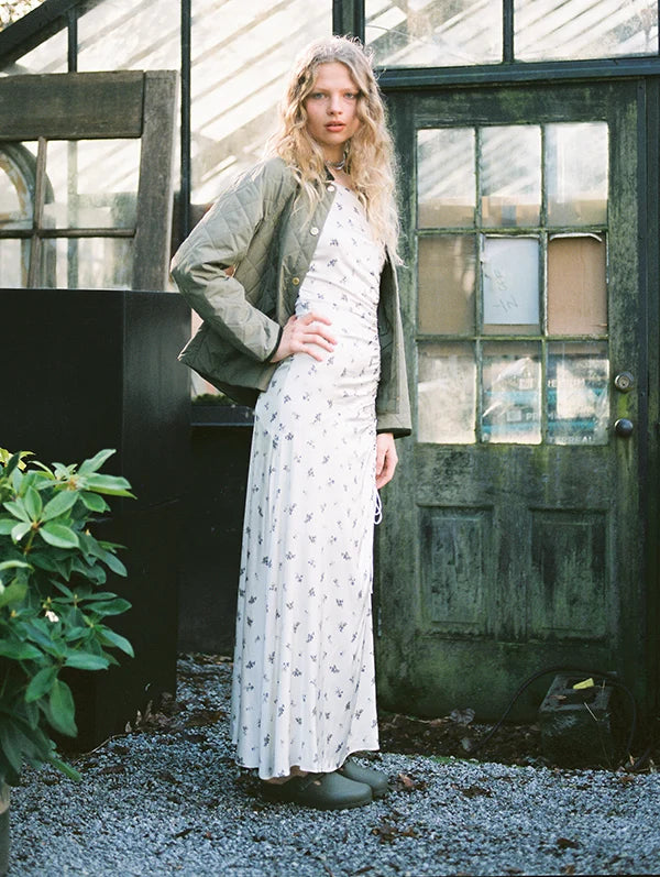 Model in floral dress and jacket posing in a greenhouse, surrounded by greenery and natural light.