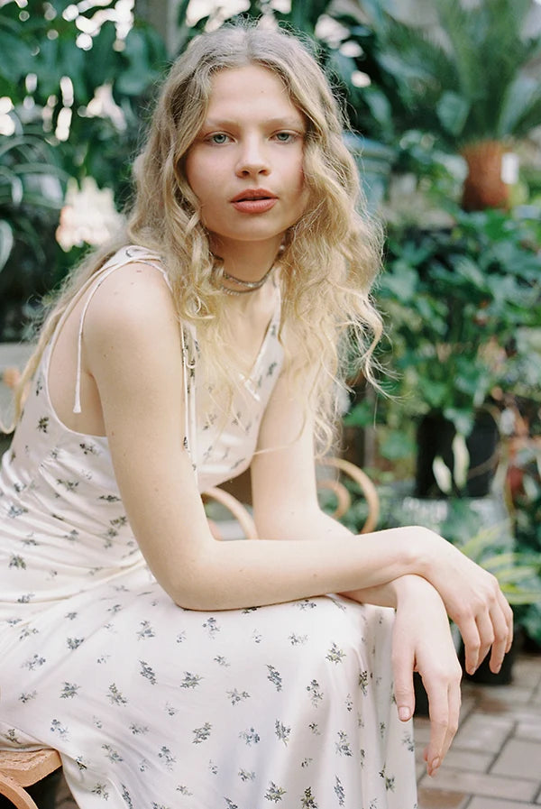 Young woman with long wavy hair in a floral dress seated in a lush green garden setting.